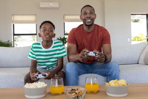 African American father and son playing video games on couch with snacks and drinks