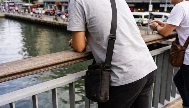 Leaning man in grey tee gazing over urban canal from railing with black crossbody bag