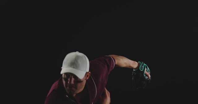 An athlete dressed in a maroon uniform and white cap is winding up to throw a pitch against a black background. The green-gloved hand is prominently highlighted, showcasing determination and action. This can be used in articles about sports, determination, team sports, or promotional material for baseball events.