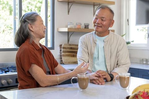 Asian senior couple holding hands at kitchen island having warm conversation over coffee