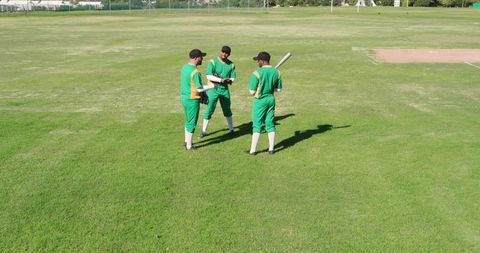 Cricket teammates huddling on sunlit field in green uniforms holding bat