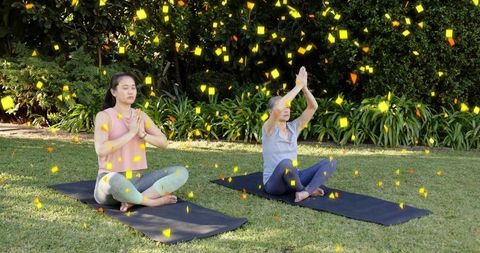 Two women practicing outdoor meditation on black mats in sunlit garden with yellow confetti