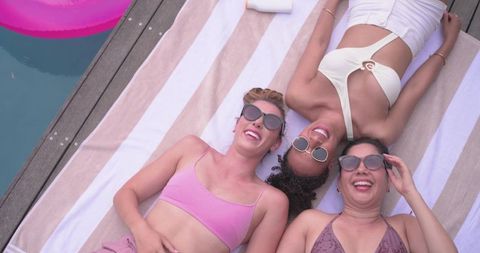 Three diverse women relaxing on pool deck laughing together in sunglasses summer vibes