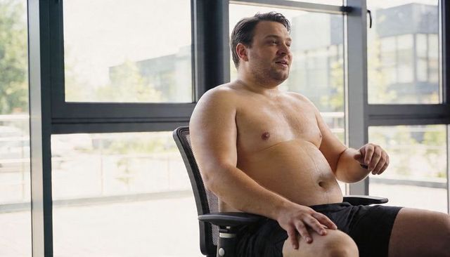 Shirtless man sitting in office chair gesturing near large windows in modern office lounge