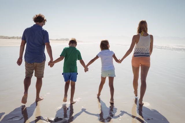 Rear view of family holding hands while walking together on shore at beach during sunny day