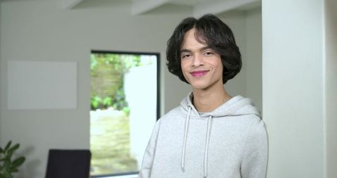 Relaxed young man leaning against pillar by window wearing gray hoodie and chain smiling