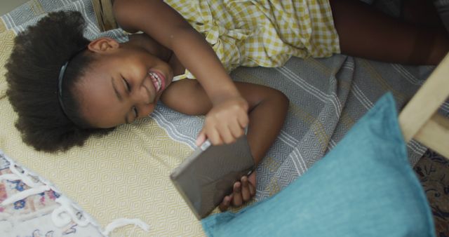 African American girl enjoying time lying on floor and using a tablet device. Suitable for topics related to childhood, education, technology use amongst children, home activities, and family leisure moments. Ideal for articles or advertisements highlighting digital learning, children's entertainment, and family friendly technology.