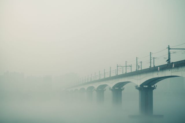 Bridge extending through a dense fog against a muted cityscape. The scene has a calm and serene atmosphere, creating a sense of mystery and tranquility. Ideal for use in articles, blogs, and marketing materials related to travel, architecture, urban lifestyle, tranquility, and weather conditions.