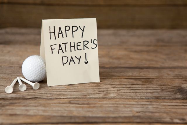 This image shows a Father's Day greeting card with a handwritten message placed next to a golf ball and golf tees on a rustic wooden table. Ideal for use in Father's Day promotions, greeting card designs, social media posts celebrating fathers, or advertisements targeting golf enthusiasts.