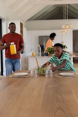 African american family arranging dining table in sunlit modern kitchen with salad and juice