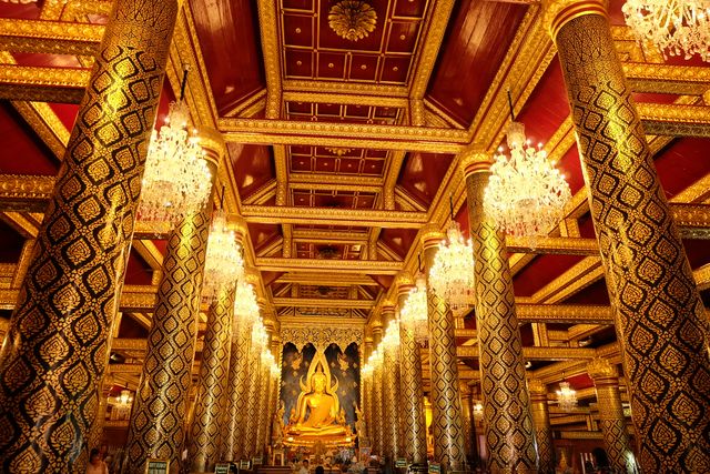 The image shows the interior of a golden temple with elaborately decorated columns and ceilings. A large Buddha statue sits prominently at the end of the hall. Chandelier lights illuminate the space, adding to the grandeur and spiritual ambiance. This image can be used in travel blogs, architecture features, cultural heritage websites, and spiritual or religious publications.