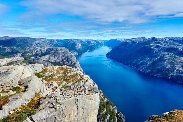 This image captures a stunning view of a Norwegian fjord surrounded by majestic cliffs and clear blue water. The scattered hikers on the cliff indicate a popular trekking destination. This image can be used for travel brochures, nature blogs, advertisements for tourism, and inspirational scenic wallpapers.