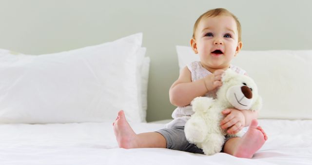 Baby sitting on a bed with white sheets, holding a teddy bear while smiling. Great for themes related to childhood, innocence, family, and home comfort. Useful for parenting blogs, baby product advertisements, and family-oriented publications.