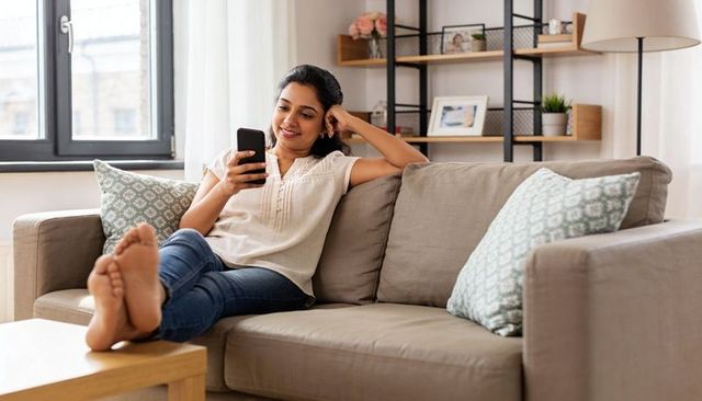 Young woman relaxing on gray sofa at home barefoot, scrolling smartphone in cozy living room