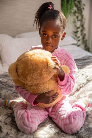 Sunlit african american girl hugging large teddy bear in pink onesie on cozy bed