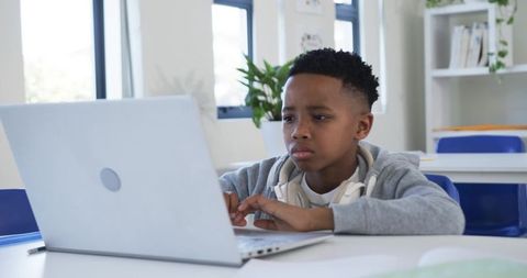 Focused schoolboy typing on laptop in bright classroom wearing headphones and grey hoodie