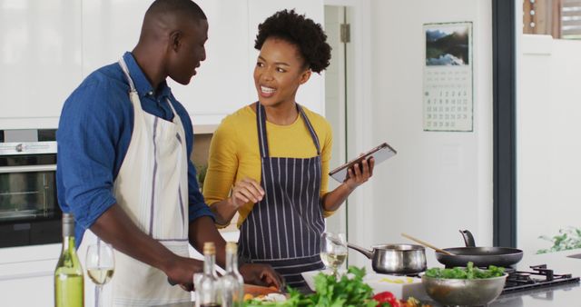 The image features a joyful African American couple cooking together in their kitchen while using a tablet. They are smiling and enjoying each other's company. Great for themes around love, relationships, healthy lifestyle, modern technology, interactive cooking, and spending quality time with loved ones. Perfect for advertisements, blog posts, websites, and social media content focused on home life, cooking tutorials, family bonding, and digital recipes.