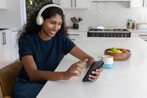 African american woman wearing headphones using smartphone at modern white kitchen island