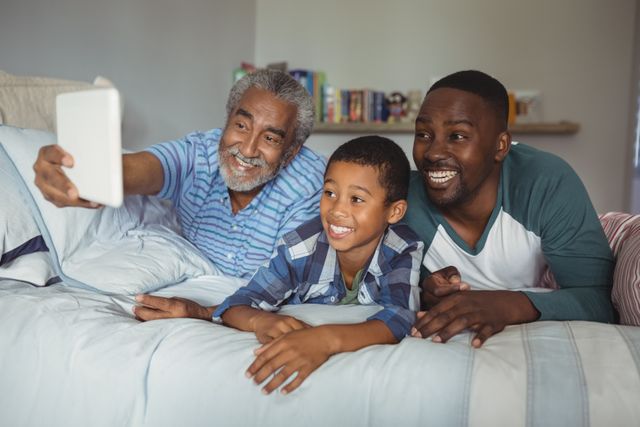 This image shows a joyful multi-generation family taking a selfie with a digital tablet while lying on a bed in a bedroom. The grandfather, father, and son are smiling and enjoying their time together. This image can be used for family-oriented content, advertisements promoting technology use in families, or articles about family bonding and togetherness.