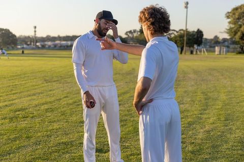 Cricket teammates discussing strategy during practice, bowler holding red ball