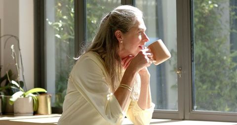 Mature Woman with Long Gray Hair Drinking Coffee by Sunlit Window