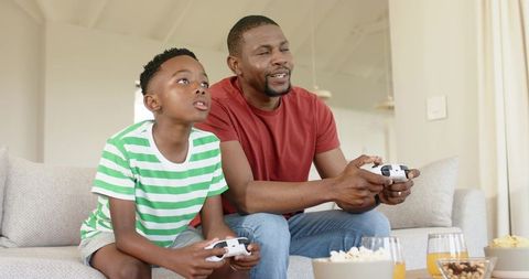 African american father and son playing video games on couch sharing popcorn
