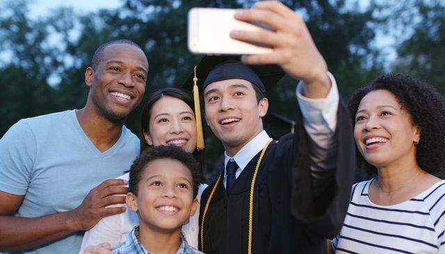 Multicultural family taking selfie with asian graduate in cap and gown on campus lawn