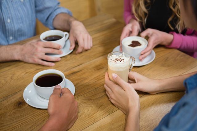 Group of friends enjoying various coffee drinks at a wooden table in a cafe. Ideal for use in articles about social gatherings, coffee culture, cafes, and friendship. Perfect for promoting coffee shops, social events, and lifestyle blogs.