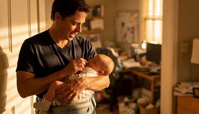 Dad holding newborn in carrier at home doorway, warm sunlight, cluttered desk behind