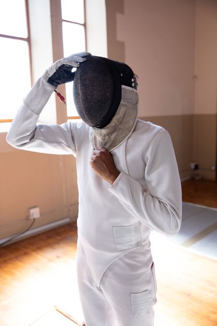 Biracial woman wearing protective fencing clothes, putting on a mask, in a gym. Sport and working out. 