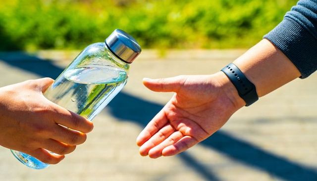 Hands passing reusable clear glass water bottle on sunlit path with fitness tracker wrist