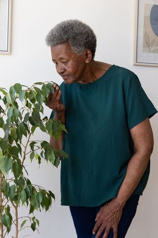 Senior african woman smelling potted plant in minimalist home interior, relaxing