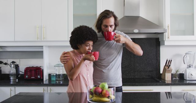 Multiethnic couple drinking coffee together in modern kitchen, embracing and enjoying morning time. Red mugs add a splash of color. Ideal for advertising home appliances, lifestyle blogs, or relationship articles.