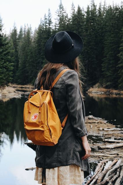 Woman hiking near a forest stream with a yellow backpack. She is wearing a black hat and an olive jacket, suggesting an adventurous spirit. This image can be used in promotional materials for travel destinations, outdoor adventure blogs, or advertisements for hiking gear and clothing.