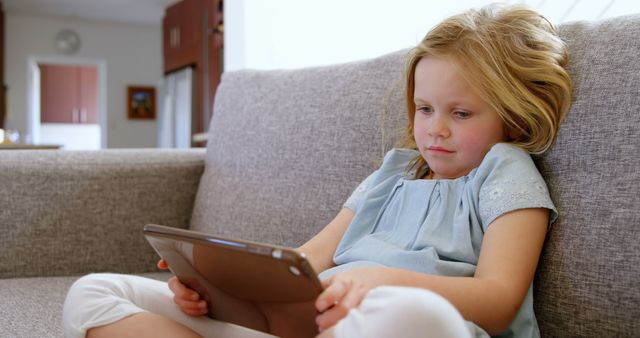 Young girl sitting on a couch using a tablet. The setting is at home with cozy, casual surroundings. Suitable for themes related to modern childhood, technology in education, family life, or relaxed home environments. Great for articles, advertisements, or educational content emphasizing relaxed, tech-savvy lifestyle.