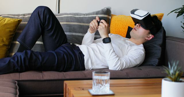 Man wearing virtual reality headset and holding gaming controller while lounging on sofa in casual, comfortable setting. Useful for topics related to technology, modern lifestyle, gaming, VR innovation, leisure activities, and interior home design.