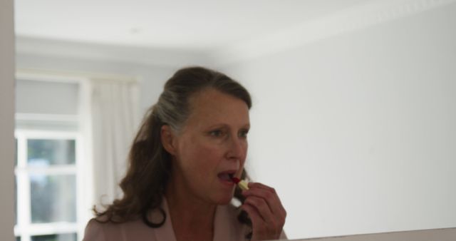 Senior caucasian woman standing in room looking in mirror and putting on lipstick. at retirement lifestyle at home.