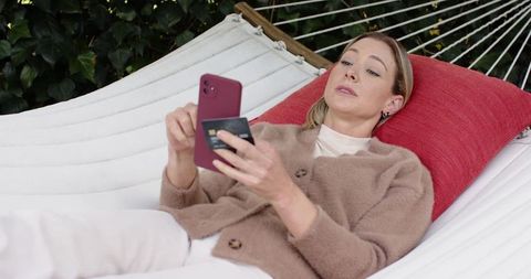 Adult woman relaxing on hammock using smartphone and credit card for mobile payment