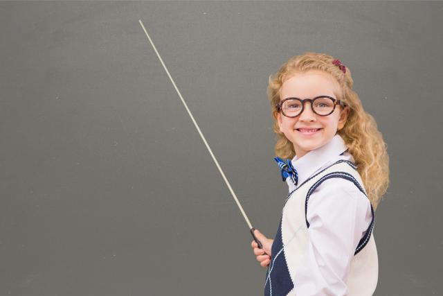 Young girl wearing glasses and bow tie, holding a pointer, and pretending to teach in front of a chalkboard. Useful for education-themed content, back to school promotions, advertisements for children's learning products, or articles discussing imaginative play and childhood education.