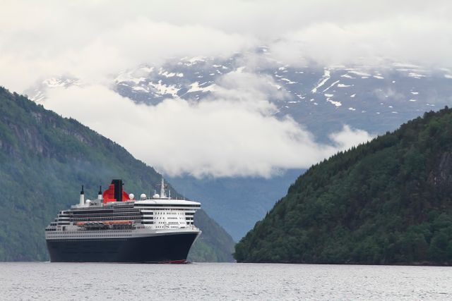 Large cruise ship sailing through a narrow fjord flanked by forested mountains and snow-capped peaks in the background, with a misty sky overhead. Ideal for travel brochures, nature-themed advertisements, or websites promoting cruises and adventure holidays.