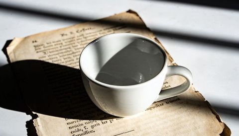White ceramic cup casting window-blind shadows on torn book pages in morning light