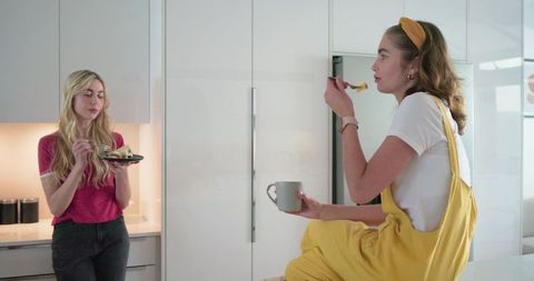 Two young women chatting over coffee and dessert in bright modern kitchen, relaxed morning