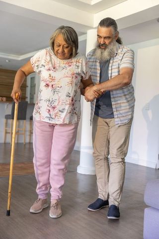 Senior couple practicing cane walking at home, caregiver assisting mobility and balance