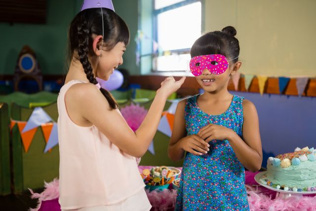 Two children are interacting at a birthday party. One child is wearing a pink mask while the other is smiling. The background features colorful decorations, a table with a cake, and party items. This image can be used for themes related to children's parties, celebrations, and joyful moments.
