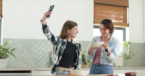 Two young women taking selfie while enjoying cooking together in sunlit modern kitchen