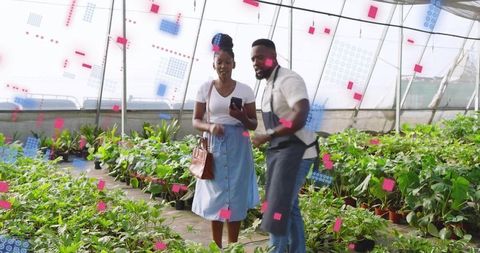 Nursery worker guiding customer through greenhouse pointing at plants and using smartphone