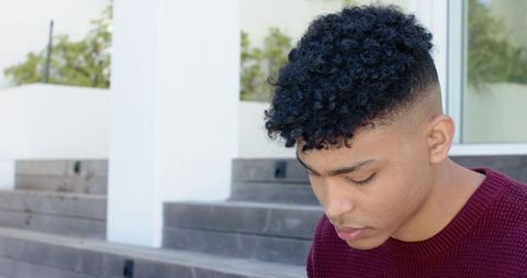 Young man sitting on concrete steps wearing burgundy sweater looking contemplative