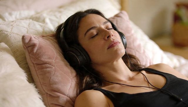 Woman resting on cozy bed listening to music with headphones on pink velvet cushion