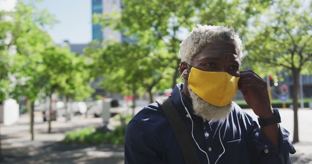 African American man with grey hair adjusting yellow face mask outside in an urban park area. He is wearing casual clothing and listening to earphones. Useful for topics on health, safety, pandemic awareness, urban living, and social distancing.
