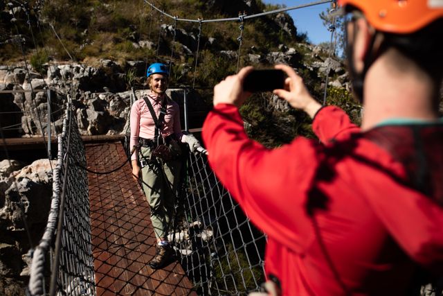 Couple enjoying an adventurous day zip lining in the mountains. The man is taking photos of the woman using a smartphone while both are wearing safety gear. Perfect for travel blogs, adventure tourism promotions, and outdoor activity advertisements.